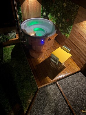 Ein Whirlpool mit einer Holzterrasse und einem kleinen Tisch, umgeben von einem Holenzaun und viel Grün.