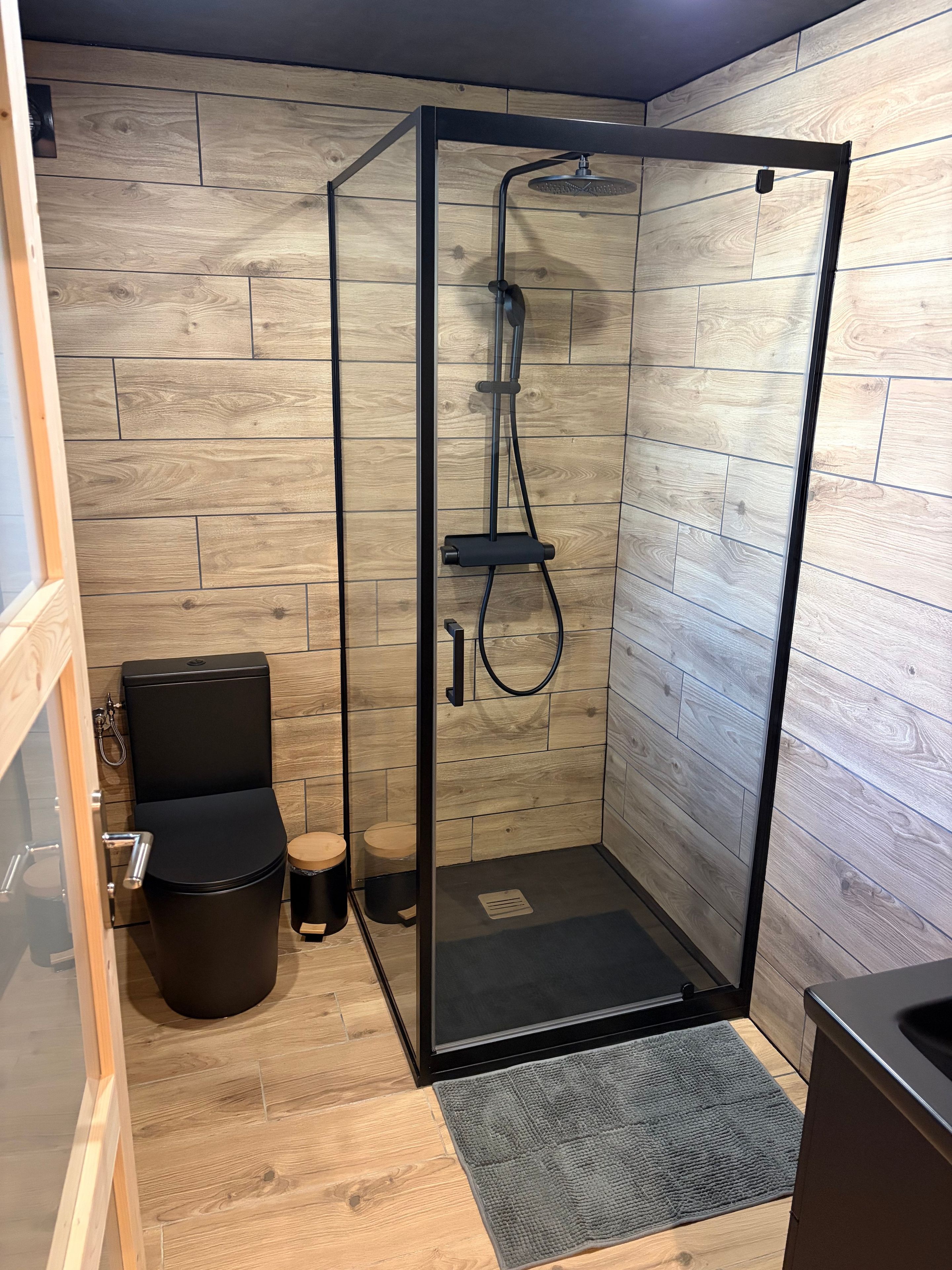 A modern bathroom with a glass-enclosed shower, a black toilet, and a wooden floor.