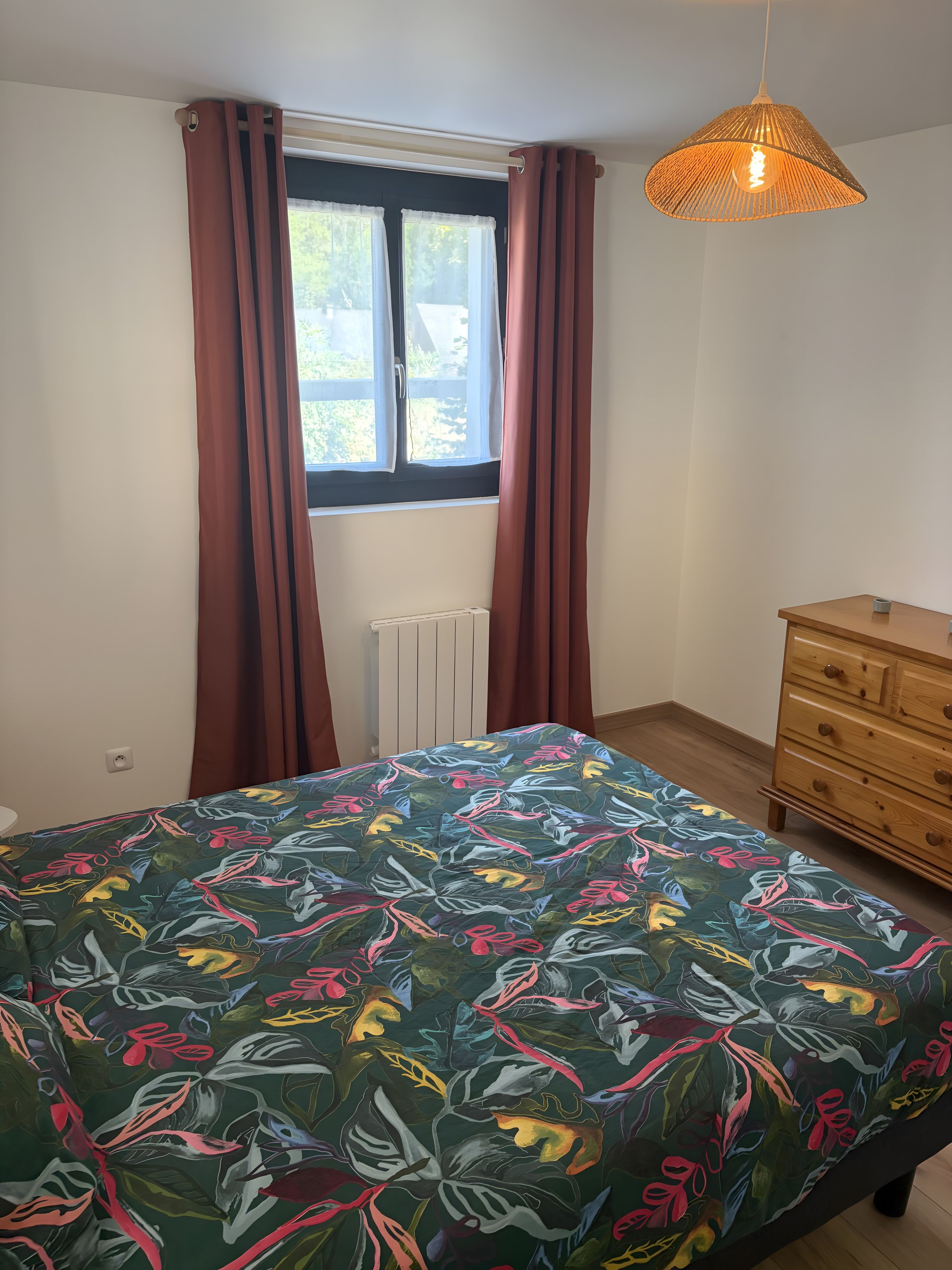 A bedroom with a bed, a dresser, a window with curtains, and a hanging light fixture.