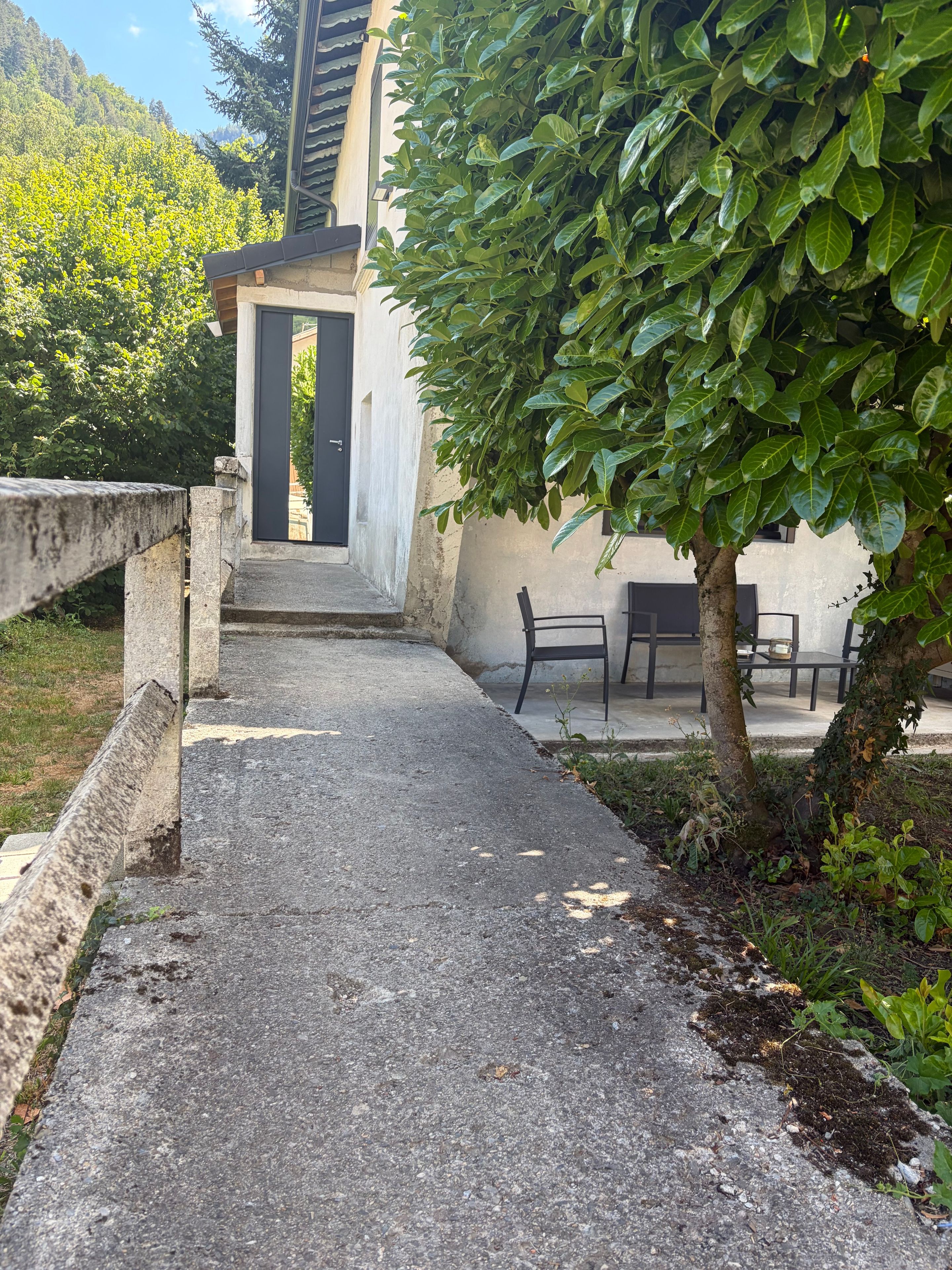 A paved pathway leading to a building with a black door, surrounded by greenery and a bench.