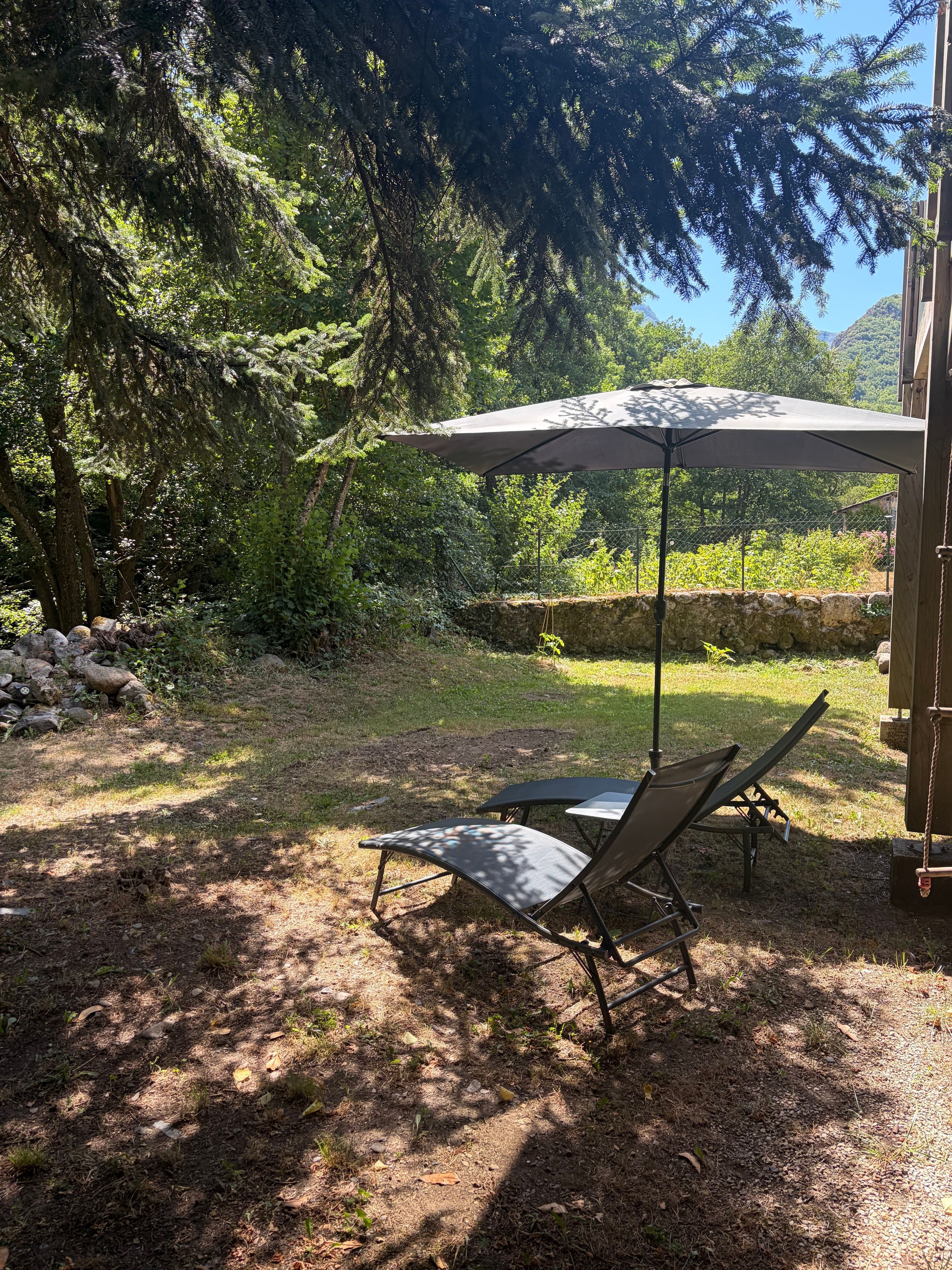 Un espace patio avec une table et deux chaises, entouré d’arbres et d’un mur de pierre.