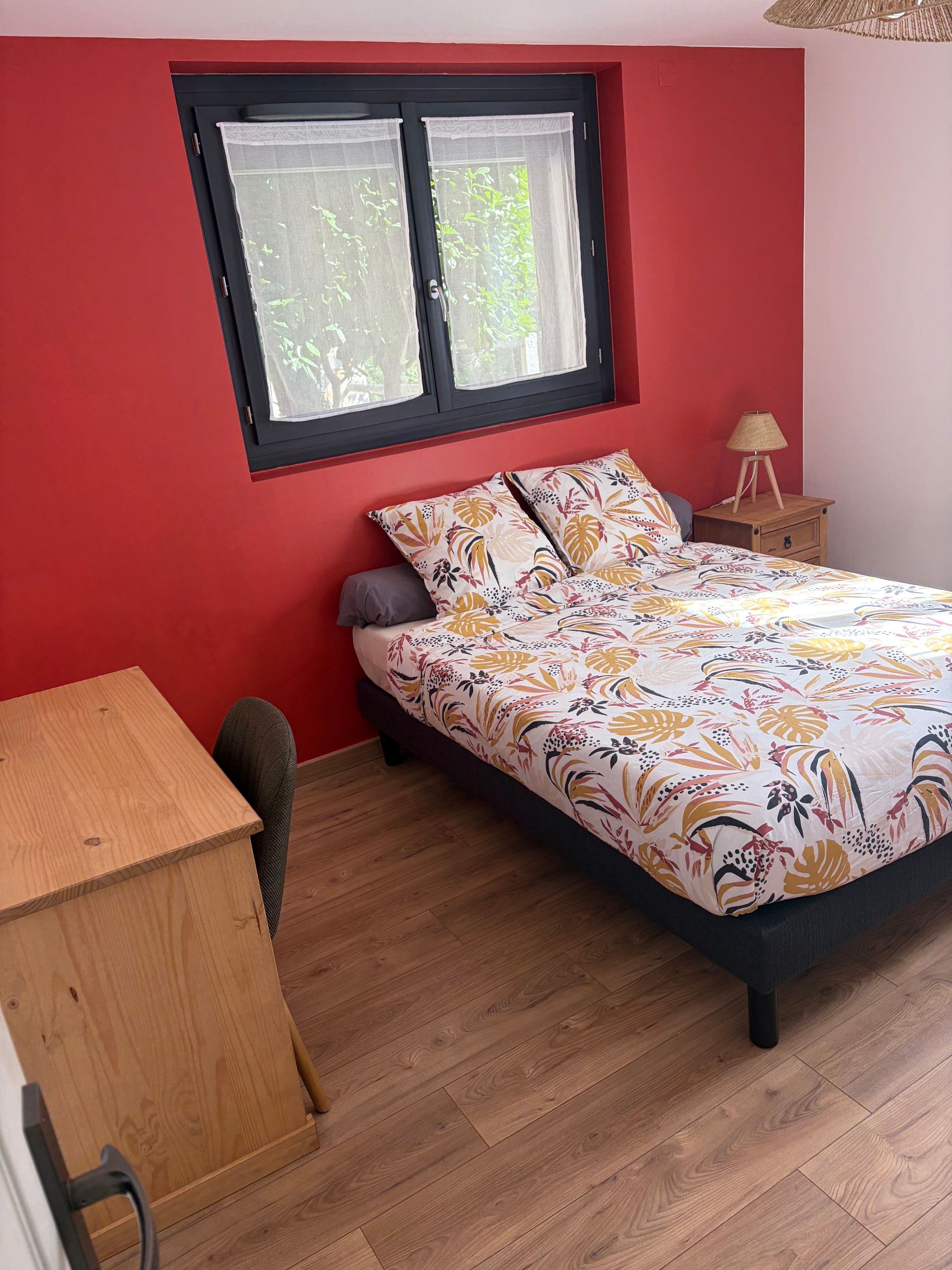 Une chambre à coucher avec un lit, un bureau en bois, une fenêtre donnant sur un paysage verdoyant et une table de chevet avec une lampe.