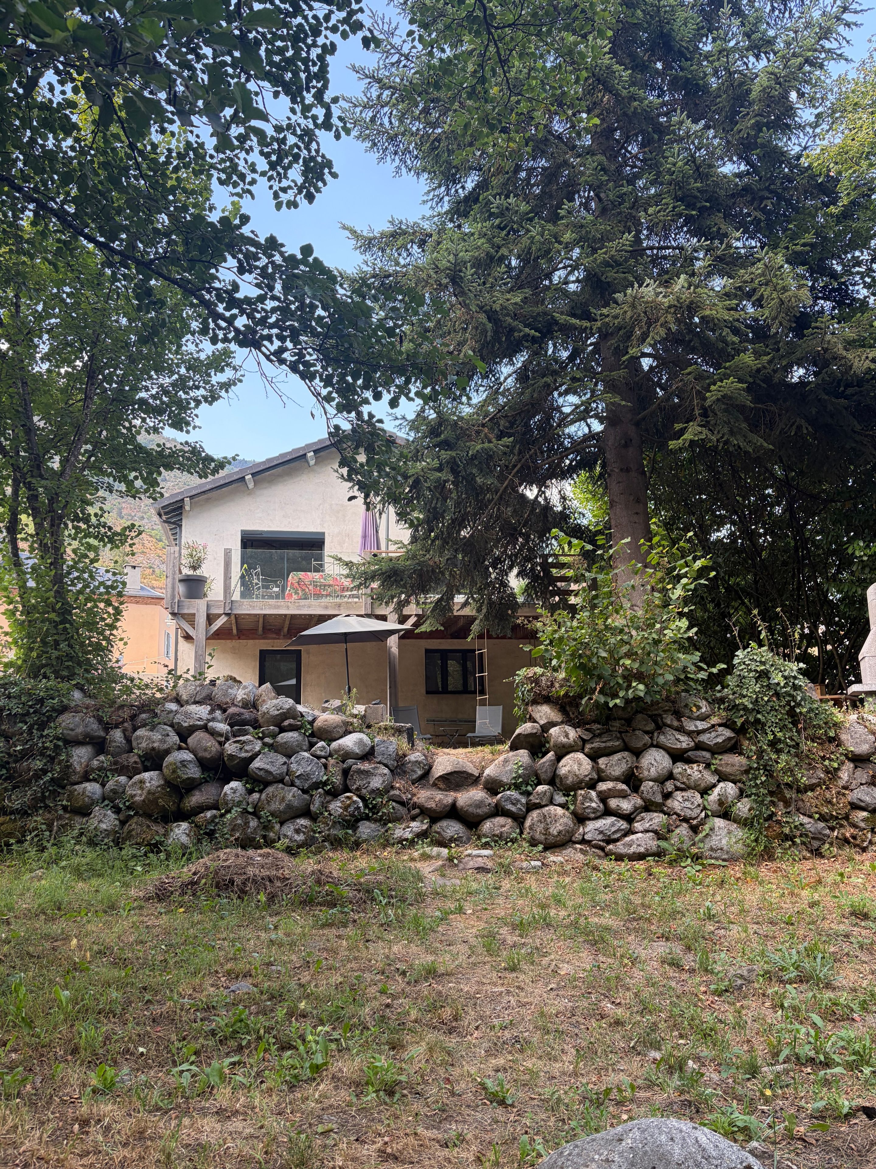 Une maison avec un mur en pierre, une terrasse et un arbre au premier plan.