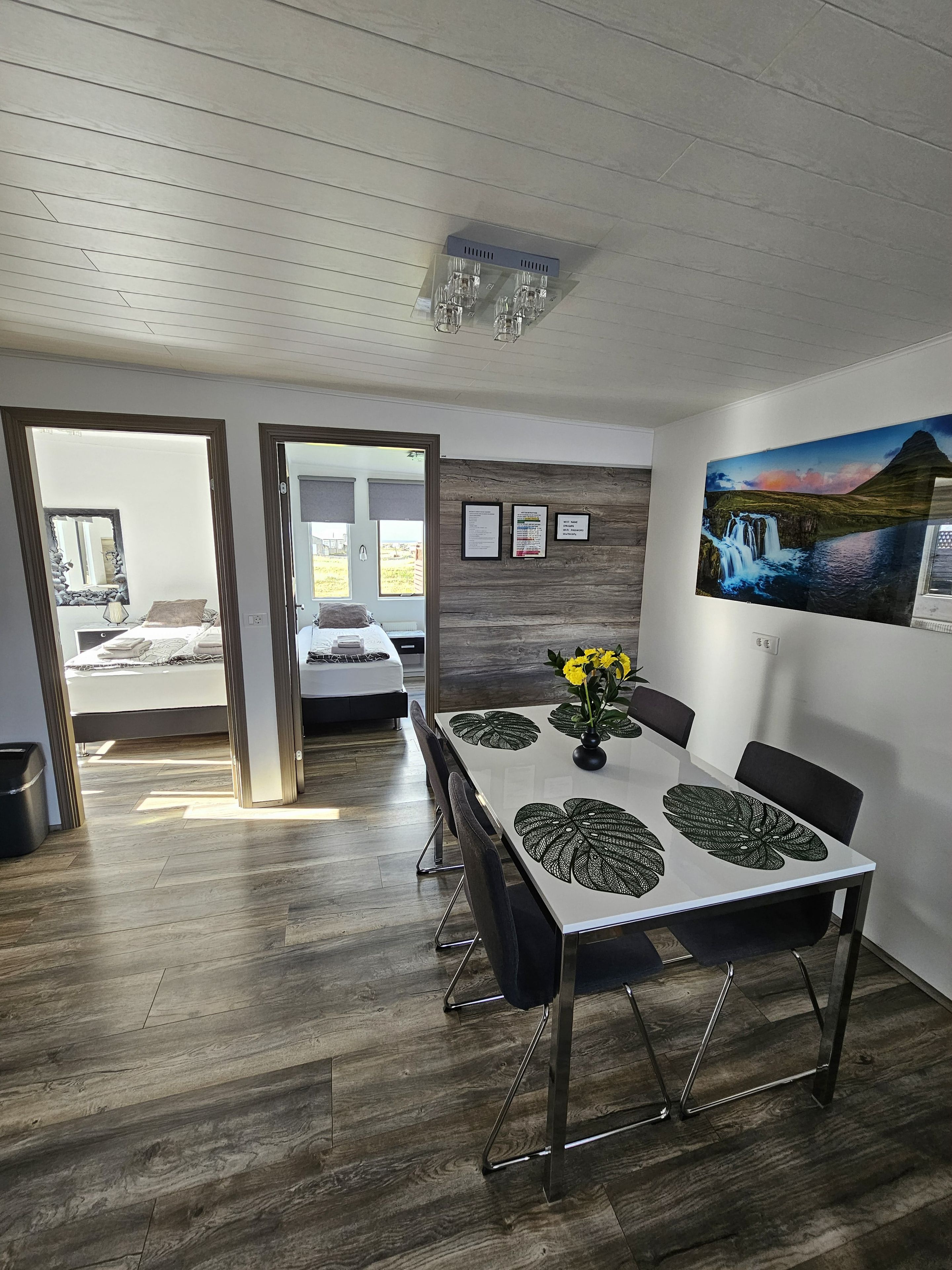 A modern dining area with a table set for four, a painting on the wall, and a view into the bedroom through an open door.