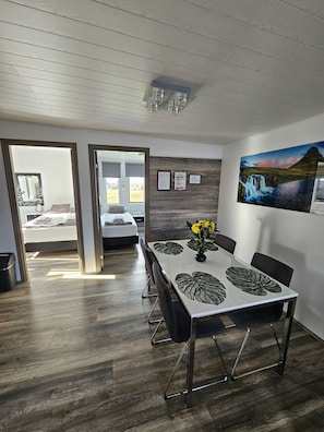 Un'area pranzo moderna con un tavolo apparecchiato per quattro persone, un quadro appeso e vista sulla camera da letto attraverso una porta aperta.