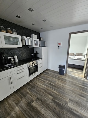 Una cucina moderna con mobili bianchi, un piano di lavoro nero e un forno a microonde. Si nota un cestino e una porta che dà su una camera da letto.
