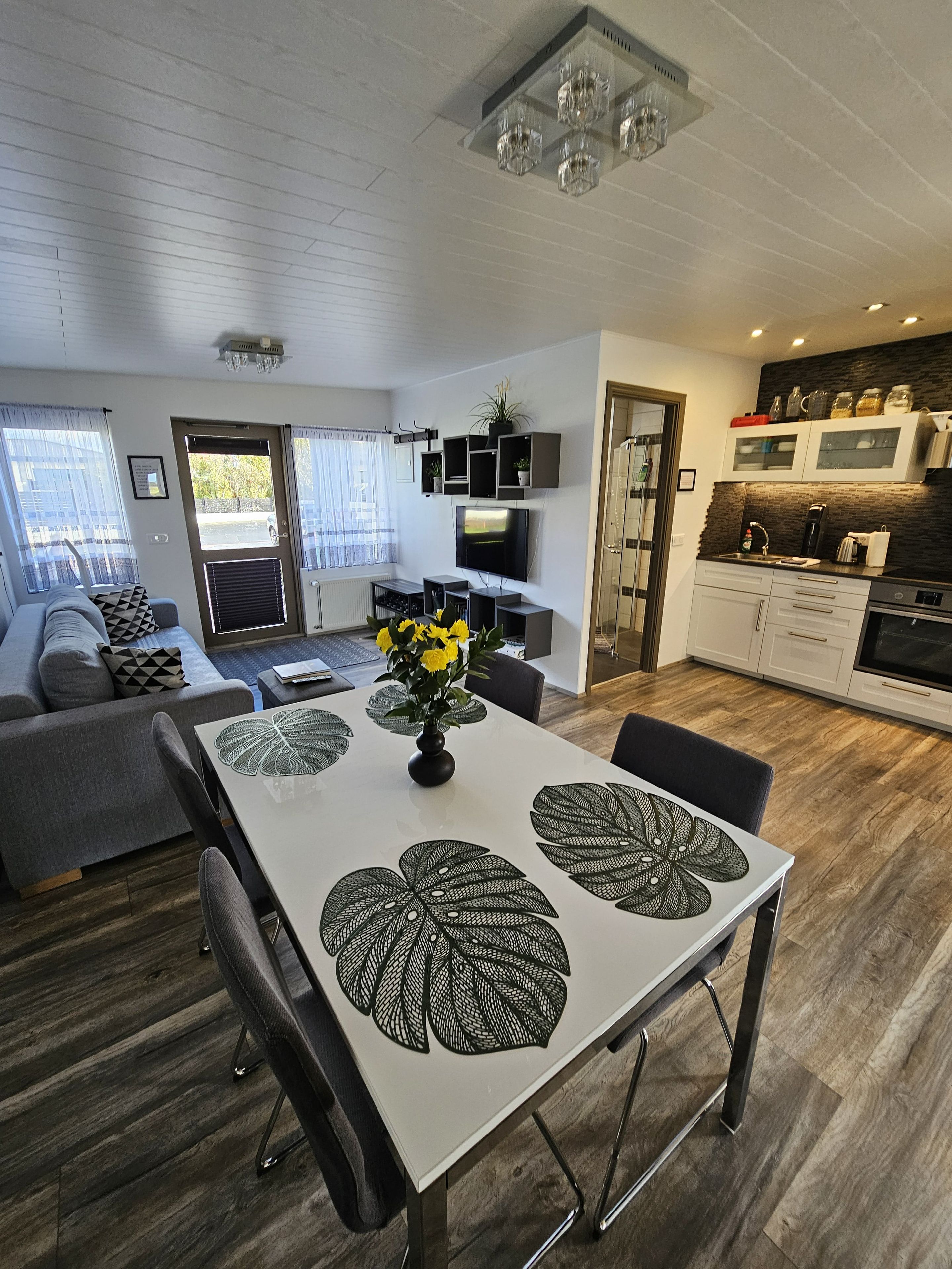 A modern kitchen with a built-in oven, a dining area with a table and chairs, and a living room with a sofa and a TV.