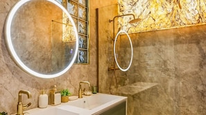 Un baño con un espejo redondo grande, cabina de ducha con mampara de vidrio y pared de mármol.