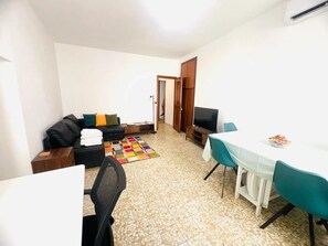 En kompakt opholdsstue med sofa, lille spisebord, fjernsyn og flisegulv.