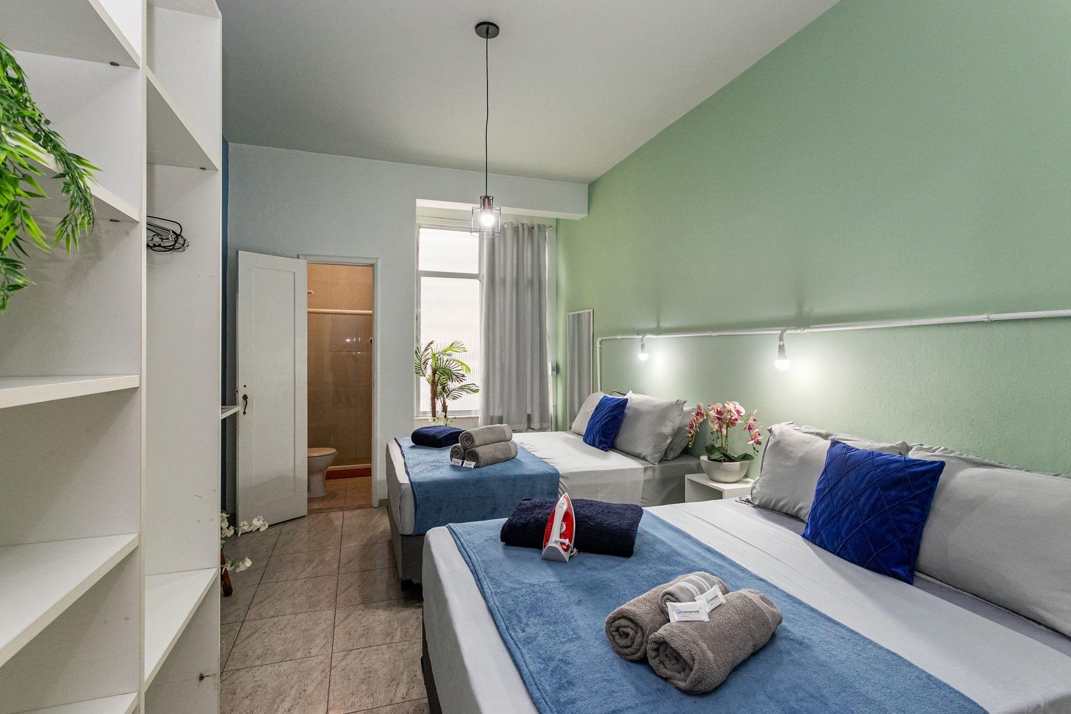 Ein Hotelzimmer mit zwei Betten, einem großen Fenster, einer Pflanze und einem durchgehenden Türspalt sichtbaren Badezimmer.