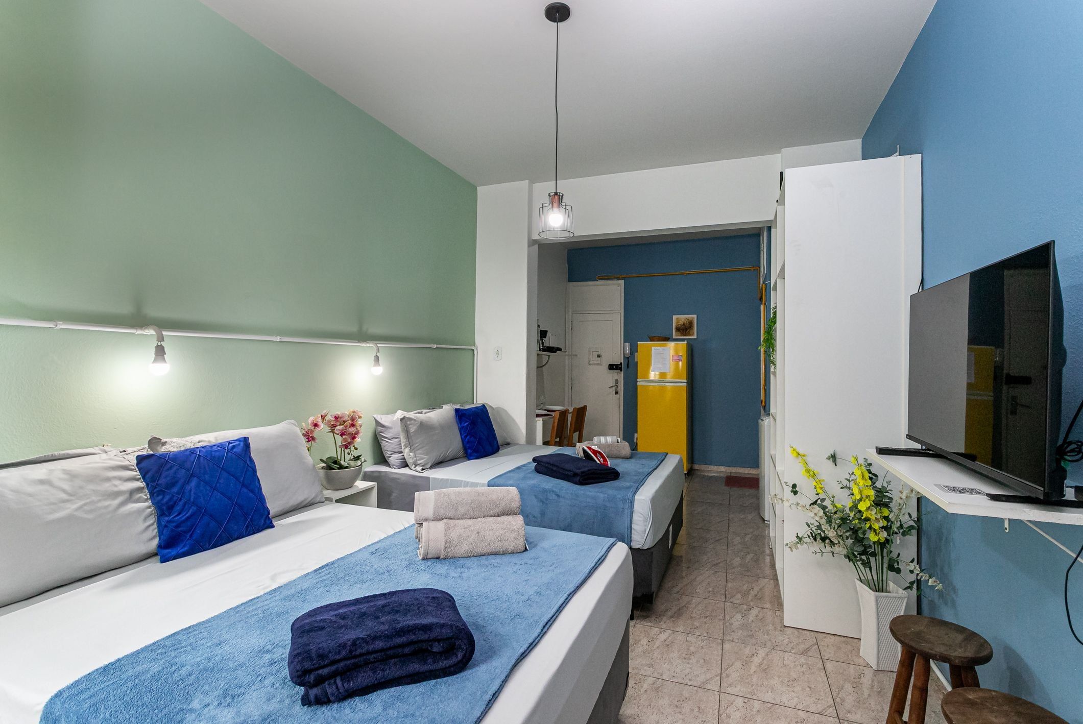 Habitación de hotel con dos camas, un escritorio con televisor, una kitchenette pequeña y un baño al fondo.