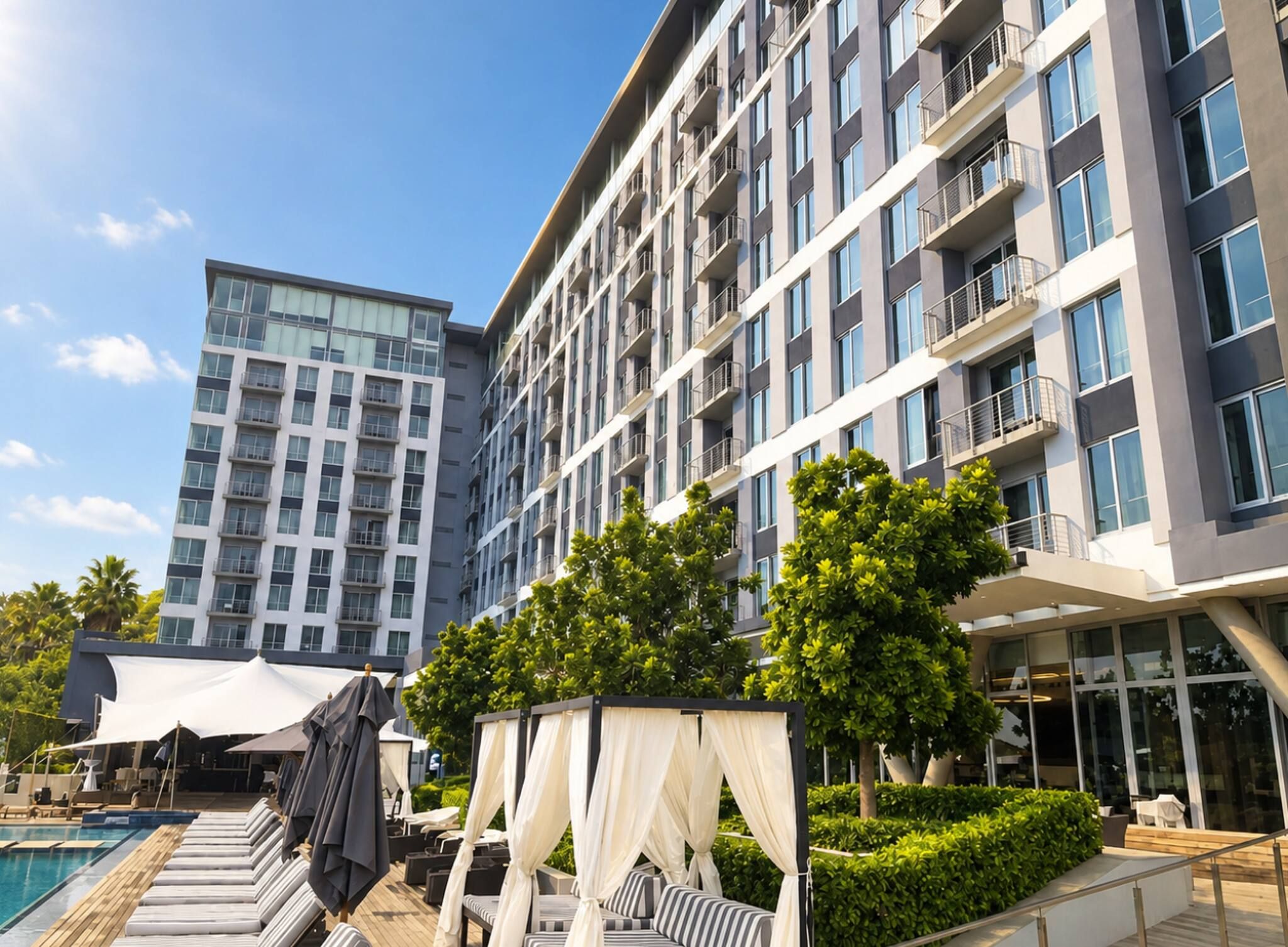 Et moderne hotel med poolområde, udendørs siddepladser og en stor bygning i baggrunden.