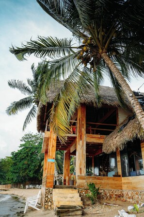 A wooden structure with a thatched roof, palm trees, and a beach in the background. - Hotel Bahía Guapinol (Mariscos)