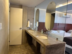 A hotel bathroom with two sinks, a mirror, and a bed visible through the glass partition. - Hotel Bahía Guapinol (Mariscos)