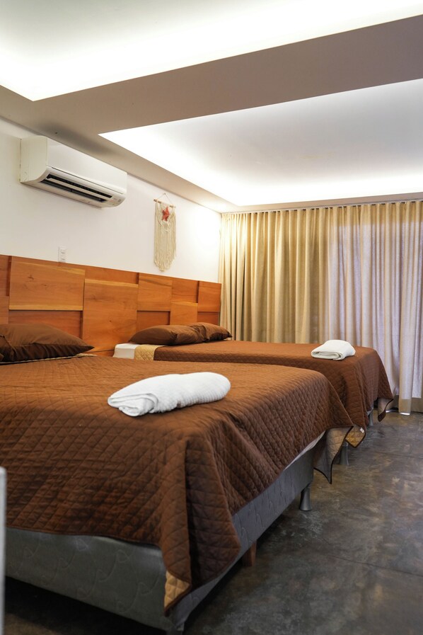 Una habitación de hotel con dos camas, un cabecero de madera, un equipo de aire acondicionado, una decoración de macramé en la pared y una cortina.