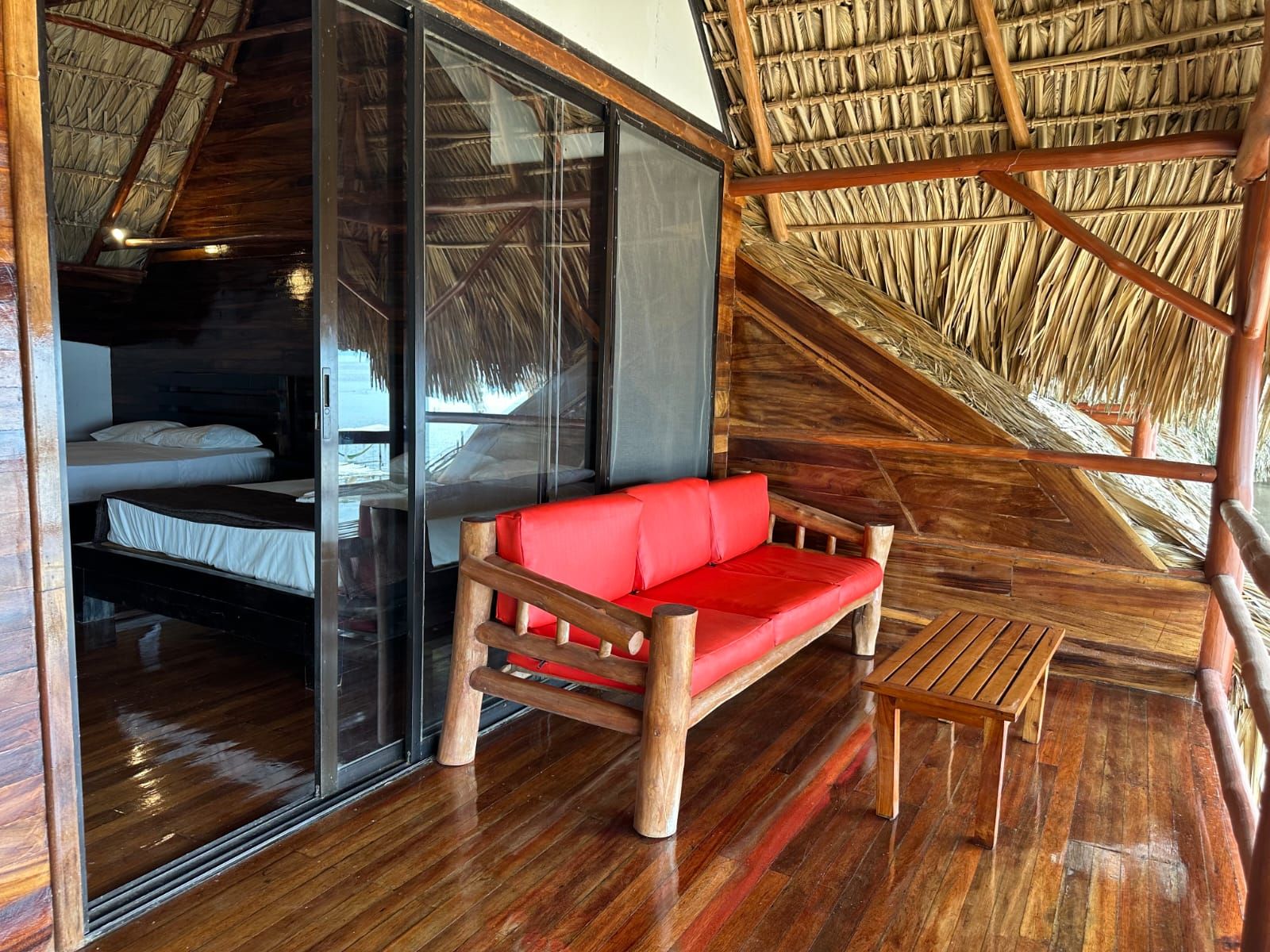 Una habitación con piso de madera, un sofá rojo, un banco de madera y un techo de paja.