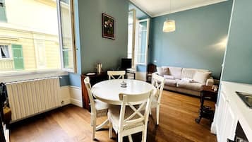 Et værelse med spisebord og stole, en sofa, en radiator og et vindue med udsigt til bygninger.
