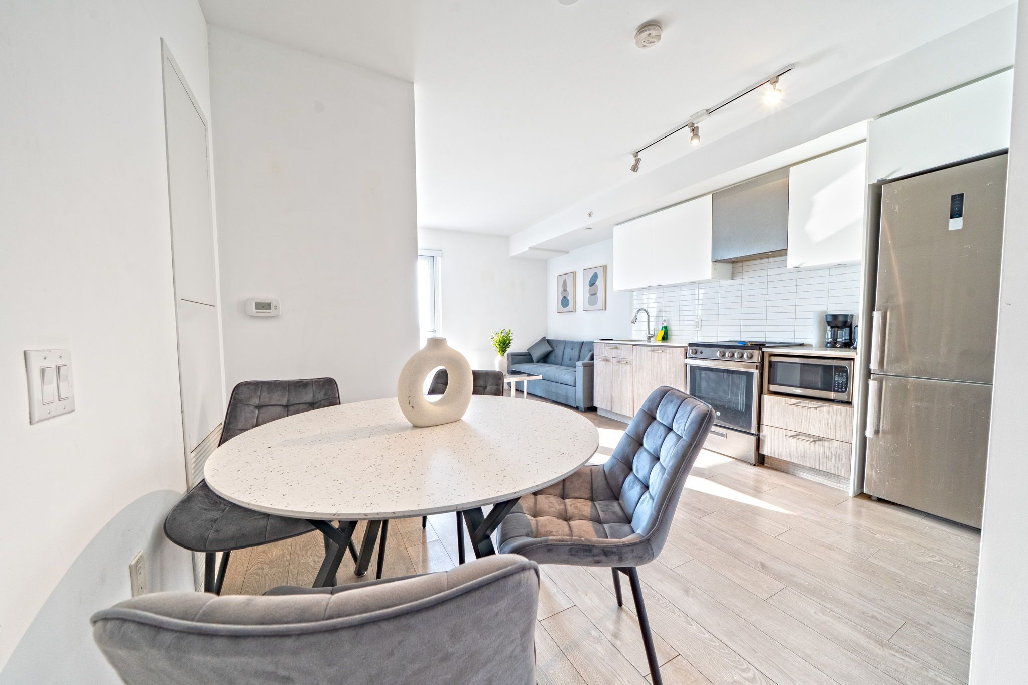A modern kitchen with a central island, a dining area with a round table and chairs, and a visible thermostat on the wall.