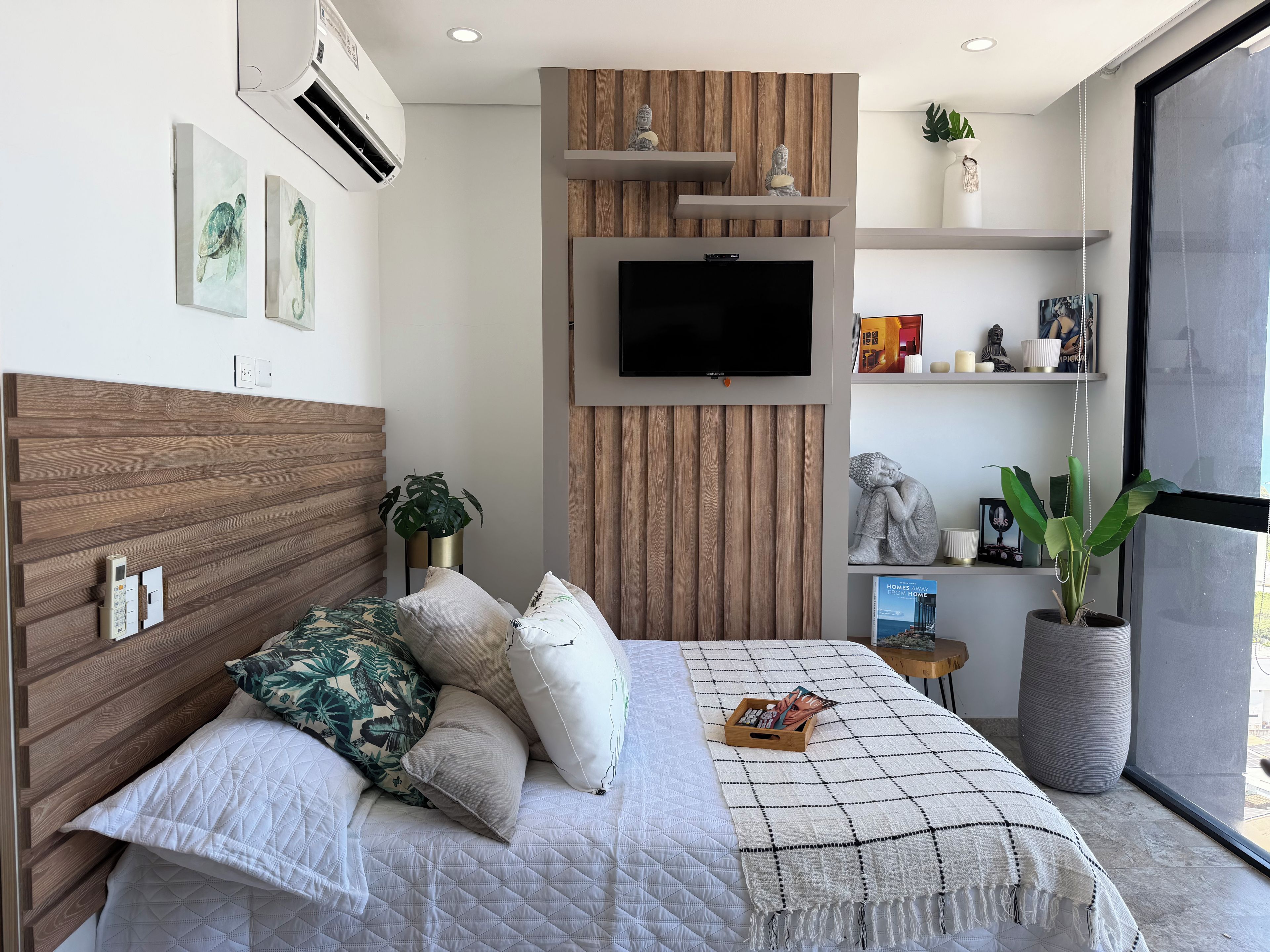 Une chambre à coucher moderne avec une tête de lit en bois, un lit agrémenté de plusieurs oreillers, un téléviseur fixé au mur et une grande fenêtre.