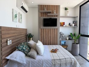 Une chambre à coucher moderne avec une tête de lit en bois, un lit agrémenté de plusieurs oreillers, un téléviseur fixé au mur et une grande fenêtre.