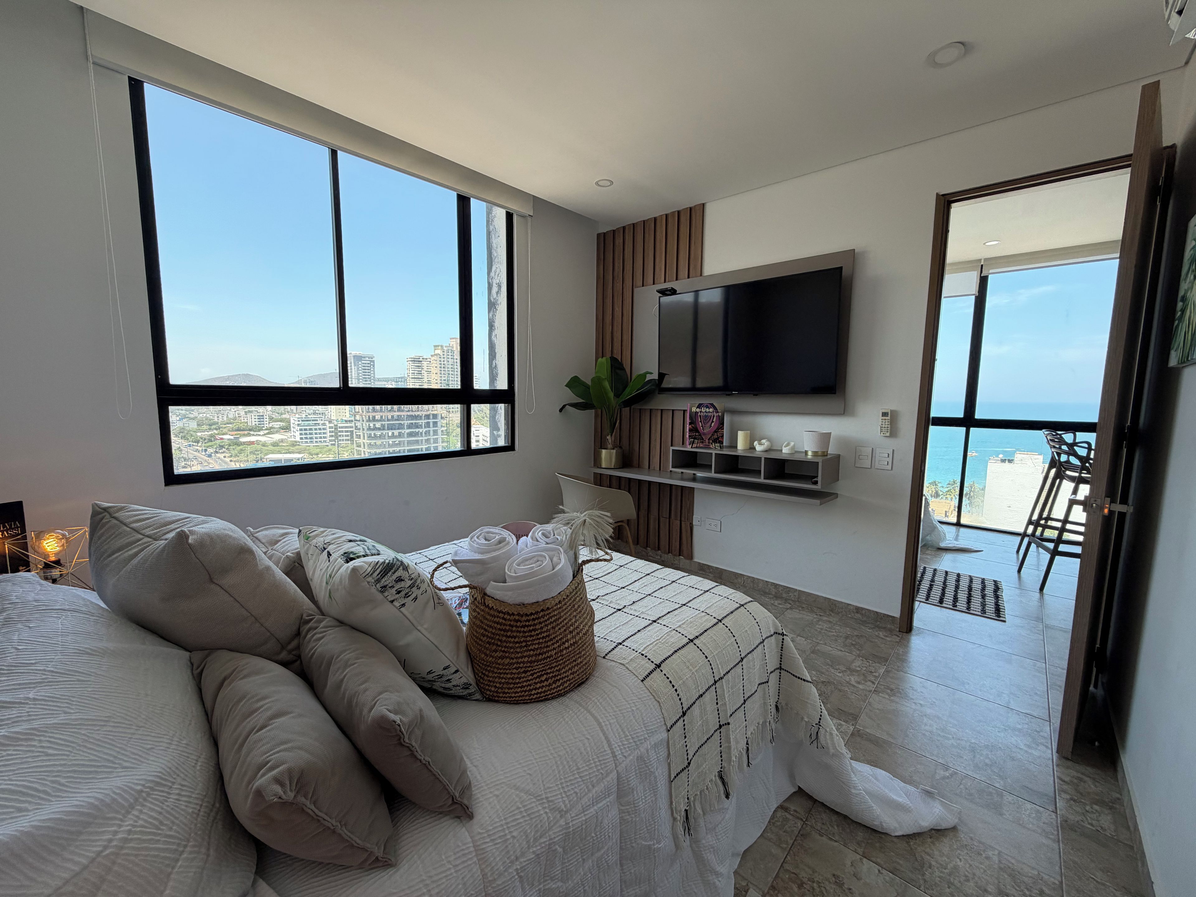 Une chambre moderne avec une grande fenêtre, un lit avec des serviettes pliées, un téléviseur fixé au mur et un balcon avec des chaises.