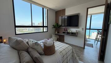 Une chambre moderne avec une grande fenêtre, un lit avec des serviettes pliées, un téléviseur fixé au mur et un balcon avec des chaises.