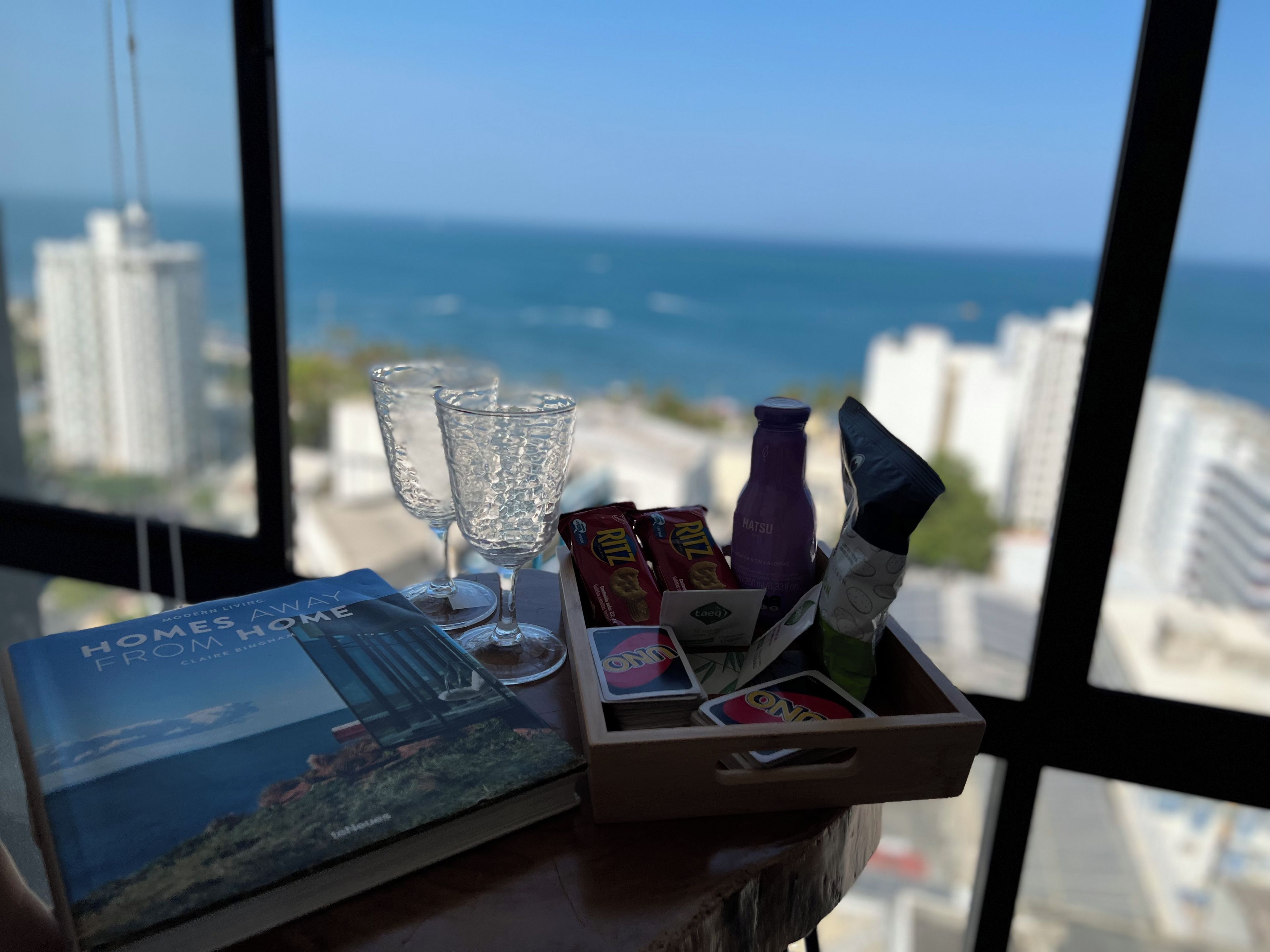 Une table avec un livre, des lunettes, des en-cas et une bouteille, avec vue sur la mer et les bâtiments.