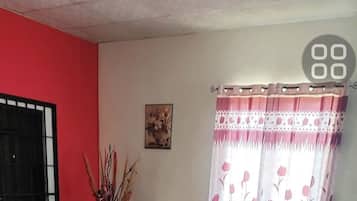 Un saló amb un sofà, una taula de centre, una cadira, un vas amb plantes seques i una finestra amb cortines florals.