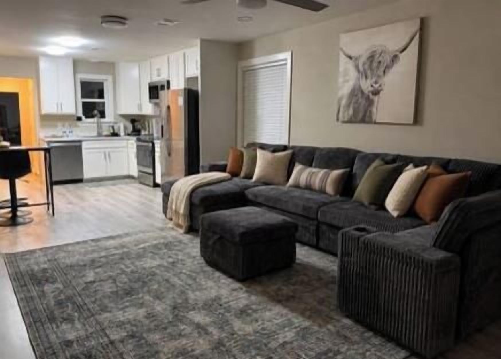 A living room with a sectional sofa, ottoman, and a rug. There is a kitchen area in the background.