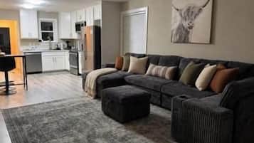 A living room with a sectional sofa, ottoman, and a rug. There is a kitchen area in the background.