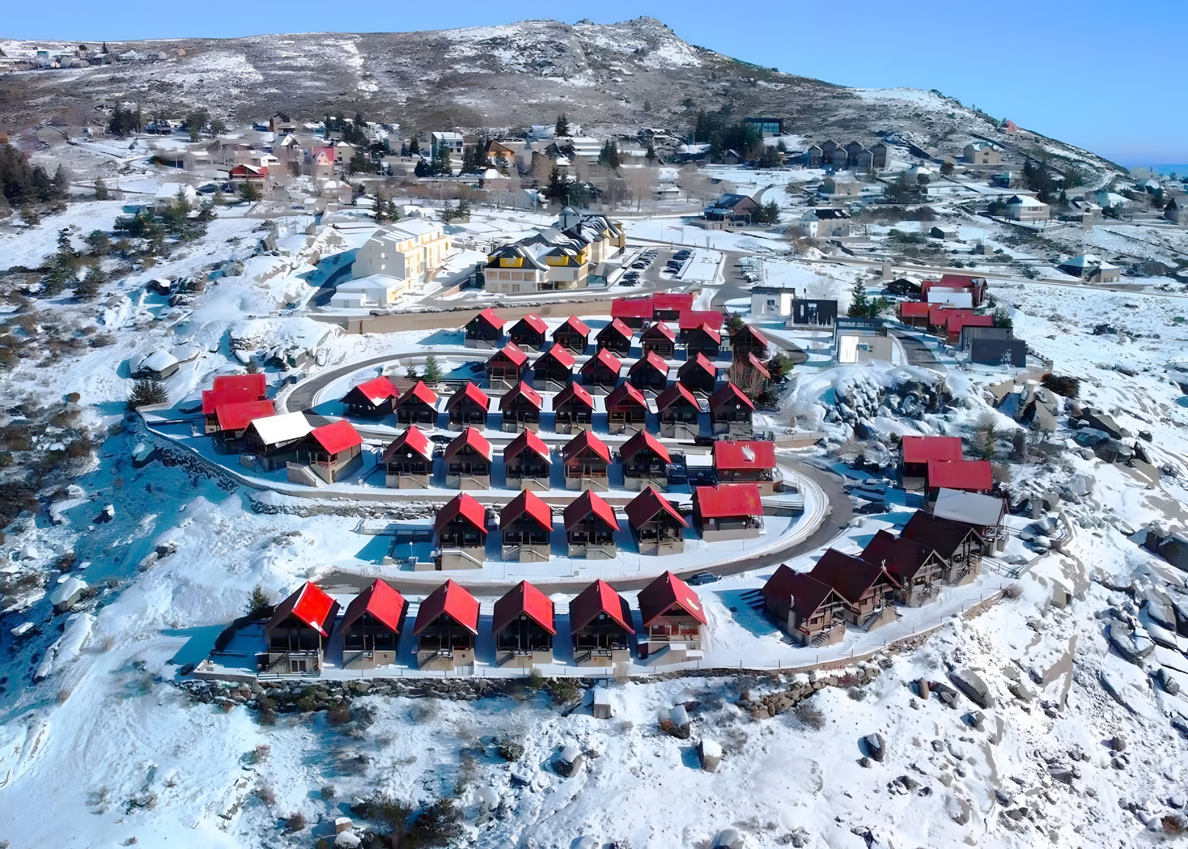 Une station de ski enneigée, avec plusieurs chalets aux toits rouges disposés en cercle autour d’une piscine centrale.