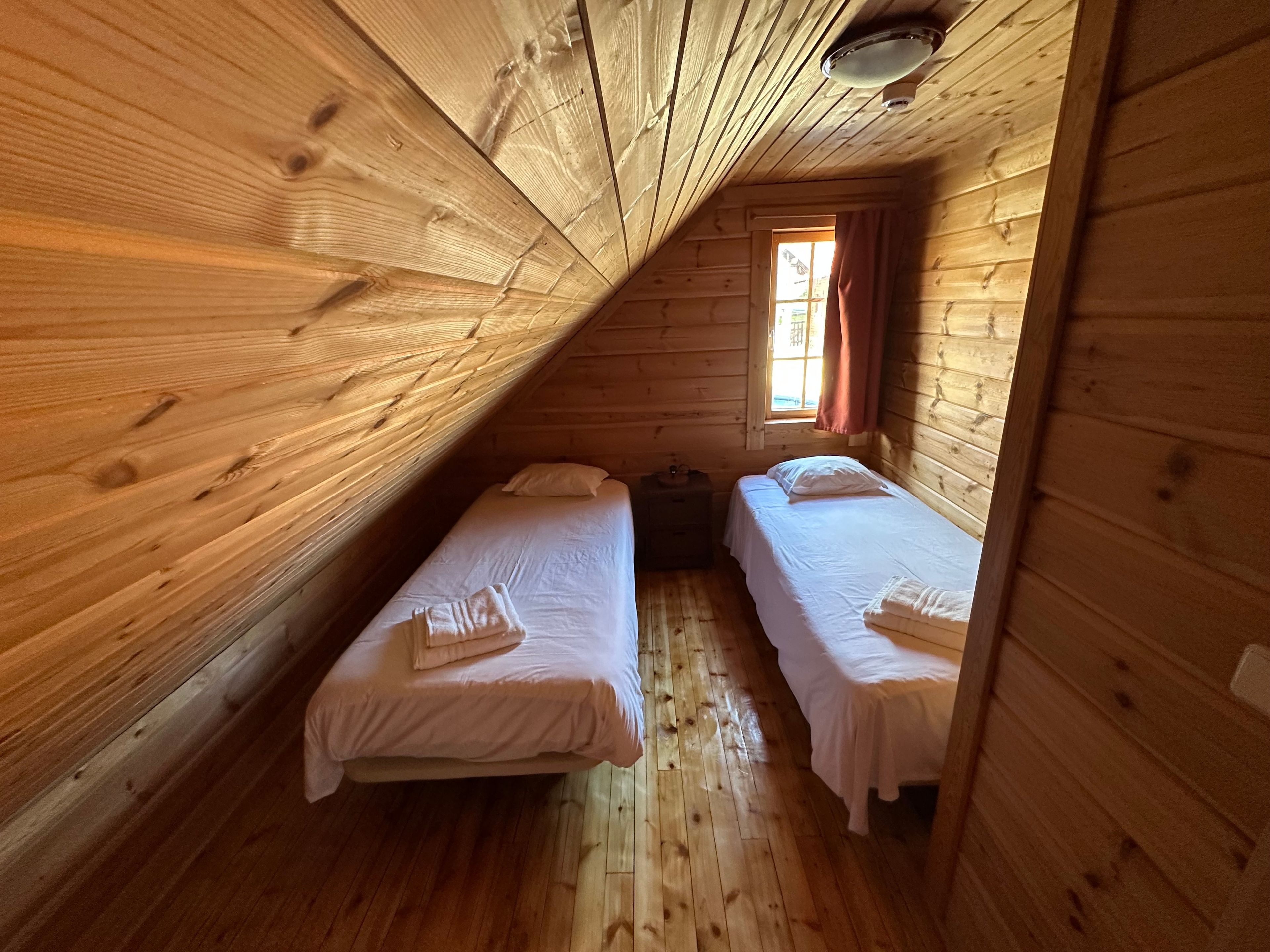 A cabin with two beds, wooden walls, a sloped ceiling, and a window with curtains.