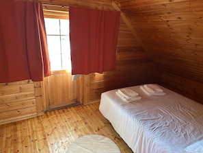 Habitación pequeña y de una sola persona con paredes de madera y techo inclinado. Tiene una cama con ropa blanca y un tapete redondo pequeño.