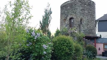 Ein steinerner Turm mit Bogenfenstern, umgeben von üppigem Grün und blühenden Pflanzen.