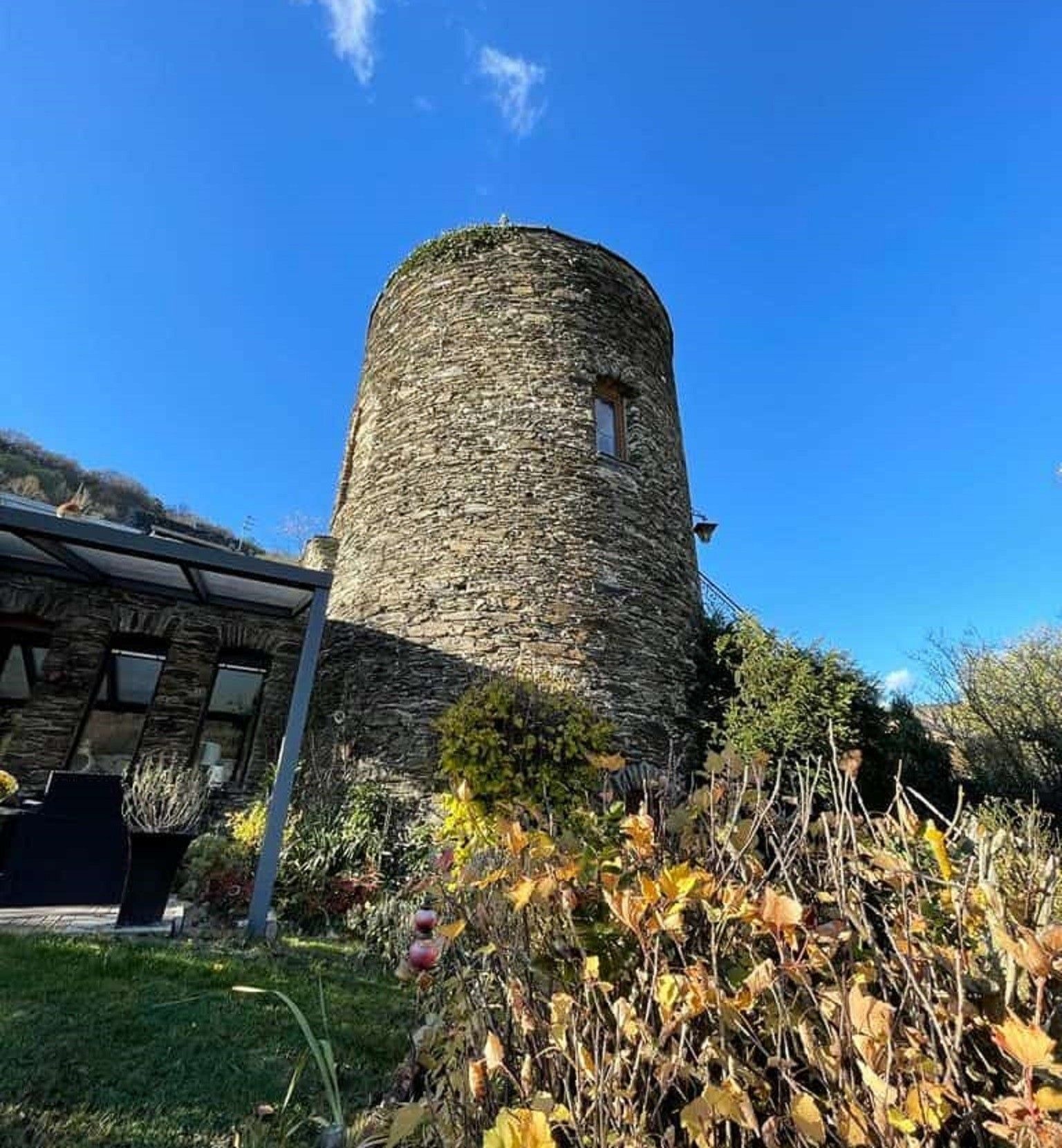 Ein steinerner Turm mit einem Fenster, umgeben von Vegetation und einem Gebäude mit geneigtem Dach.