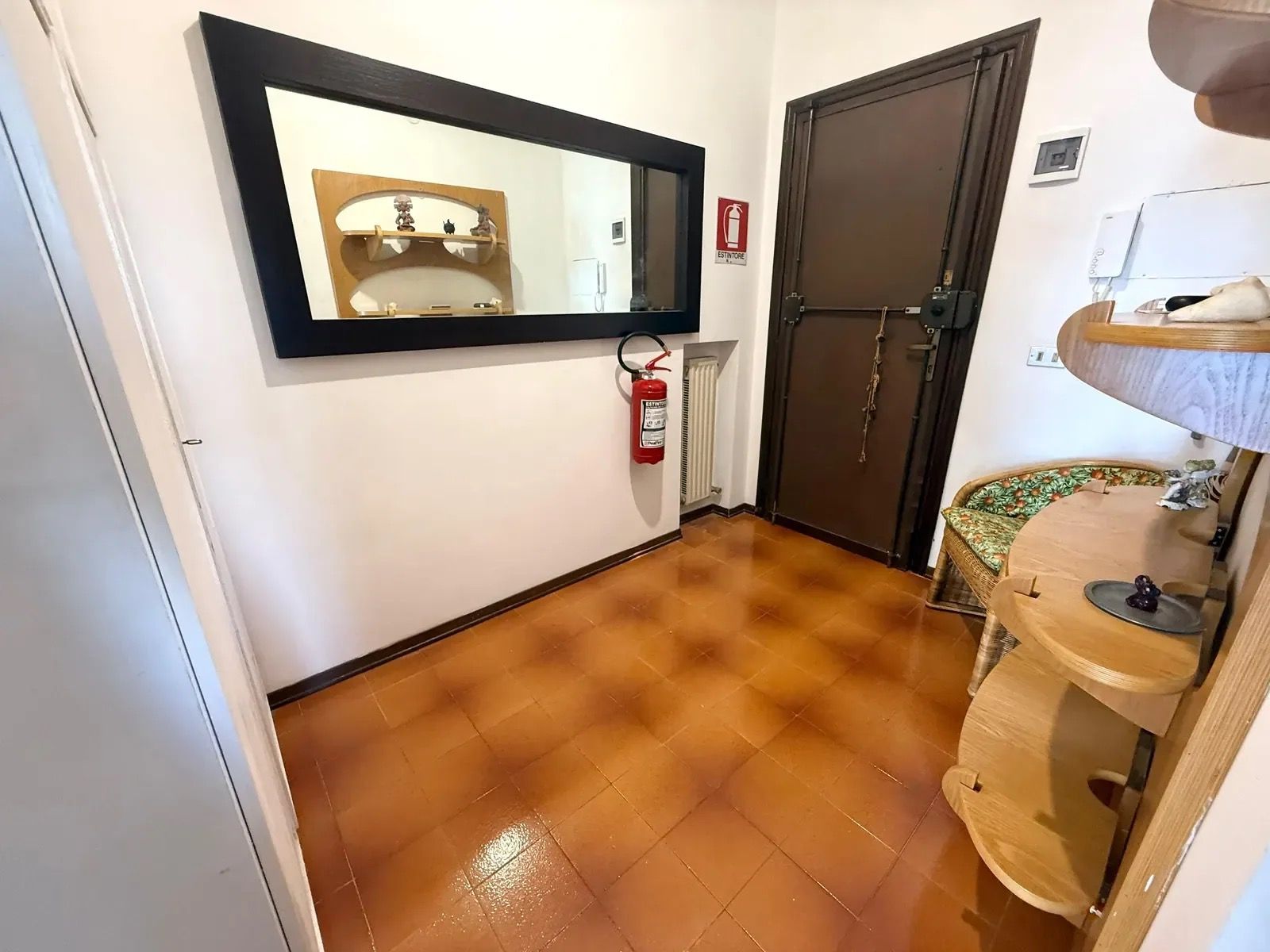 A hallway with a tiled floor, a wooden door, a mirror, a fire extinguisher, and a small sink with a towel.
