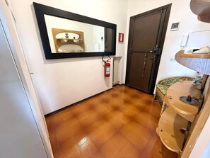 A hallway with a tiled floor, a wooden door, a mirror, a fire extinguisher, and a small sink with a towel. - New Listing (Sanremo)