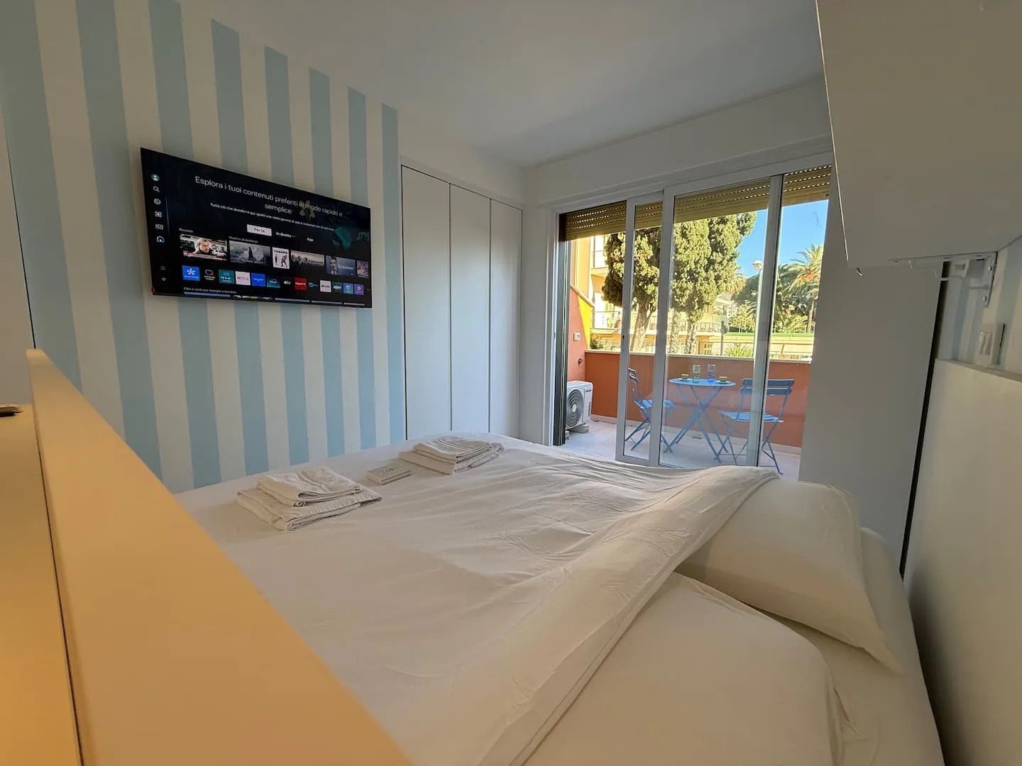 Habitación de hotel con una cama, un televisor montado en la pared y un balcón con mesa y sillas.