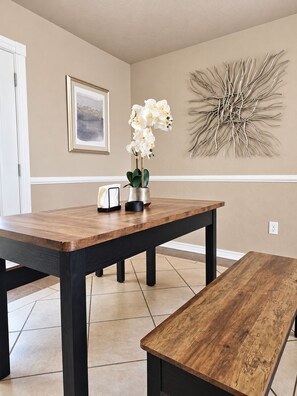 Een eetgedeelte met een houten tafel en een bank, een vaas met witte bloemen, een ingelijste foto en een kunstwerk aan de muur.