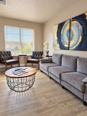Ein Wohnzimmer mit grauem Sofa, zwei Sesseln, einem Couchtisch und einem großen abstrakten Gemälde an der Wand.