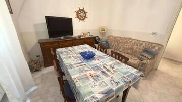 Ein Wohnzimmer mit Esstisch, bedeckt mit einer gemusterten Tischdecke, einer Couch, einem Fernseher und einer Wanduhr.