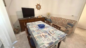 Ein Wohnzimmer mit Esstisch, bedeckt mit einer gemusterten Tischdecke, einer Couch, einem Fernseher und einer Wanduhr.