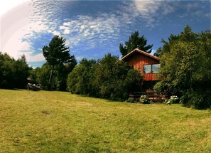 A wooden cabin with a deck, surrounded by trees and a grassy field. - New Listing (Quetroleufu)