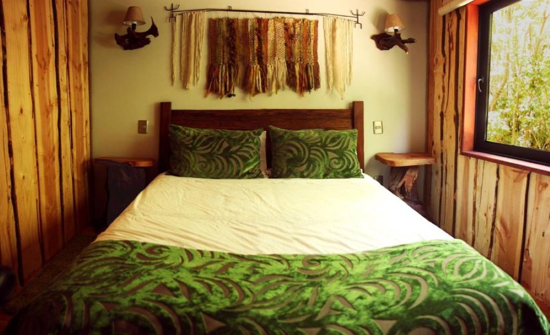 A wooden room with a bed, two wall-mounted lamps, and a window with a view of greenery.