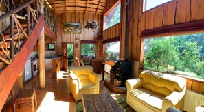Ein Interieur einer Holzhütte mit Treppe, gelben Sofas, einem Kamin und großen Fenstern mit Blick auf Grünflächen.