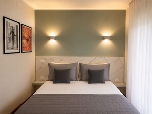 Hotellihuone, jossa on sänky, kaksi tyynyä, sängynpääty, jossa on seinään kiinnitetyt valaisimet, ja kaksi kehystettyä kuvaa seinällä.