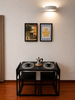 A dining area with a table set for two, a coffee maker, and two cups. There are two framed pictures on the wall, one of a café scene and another with text. - AE Nova Studio – Panoramic Views of Caserta (Caserta)