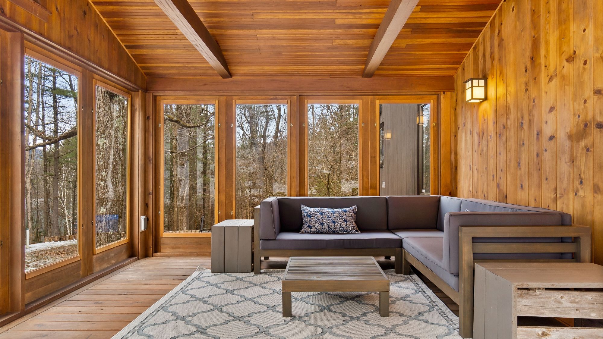 A wooden interior with a sectional sofa, a coffee table, and large windows offering a forest view.
