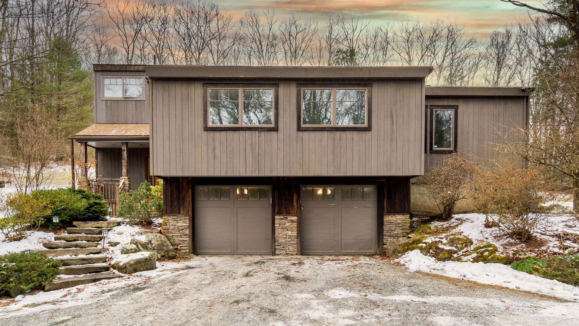 A two-story house with a garage, stone foundation, and a view of trees in the background.