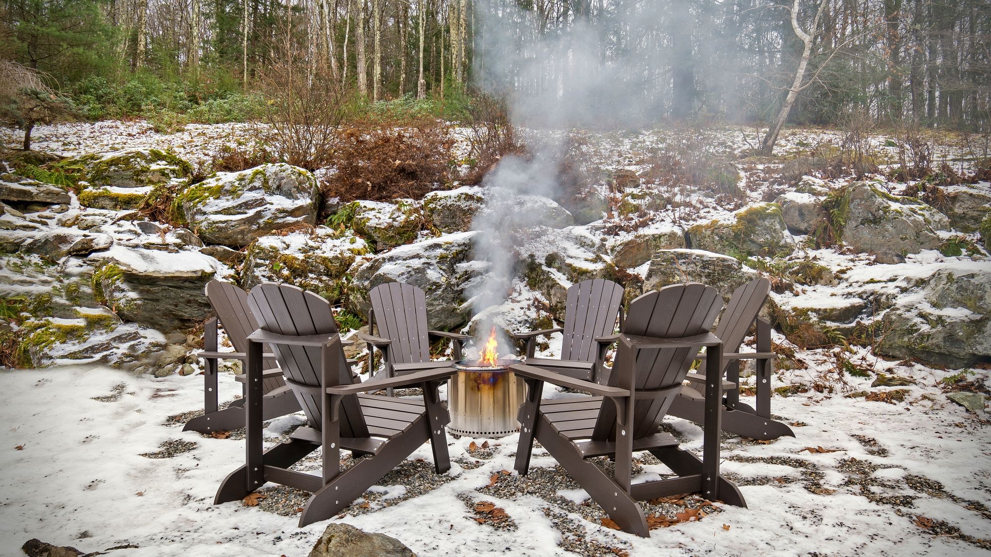 A snow-covered outdoor area with a fire pit and four Adirondack chairs arranged around it.